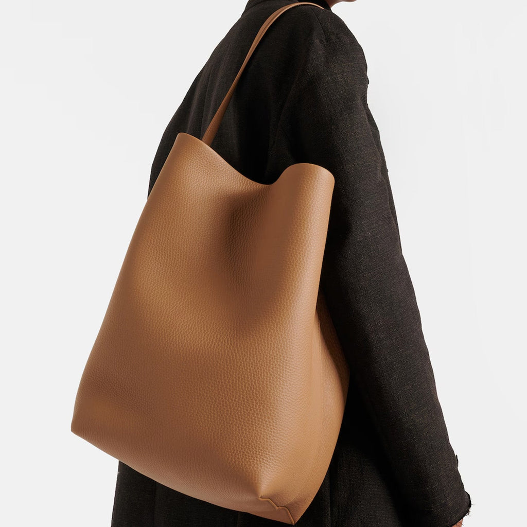 Large vertical tote bag in warm brown vegan PU leather with soft structure, minimalist design, and wide shoulder straps for everyday wear.