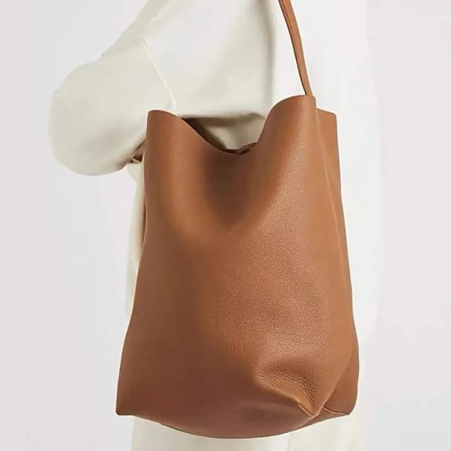 Large vertical tote bag in warm brown vegan PU leather with soft structure, minimalist design, and wide shoulder straps for everyday wear.