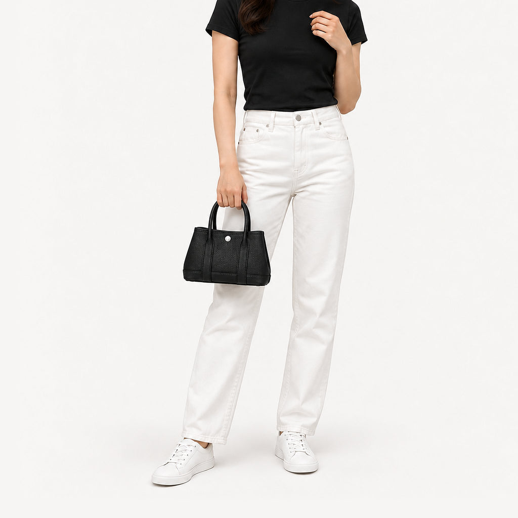 Weekday Tote Mini 23 cm open-top tote in black carried by the top handles with a black tee and white straight-leg jeans, model shot showing the 23 cm size on the body